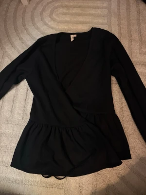 Svart omlottopp från Nelly - Säljer en svart omlottopp från Nelly med lång ärm och v-ringad hals. Toppen har en markerad midja och volang nedtill som ger en snygg siluett. Materialet är ribbat och stretchigt. Storlek S. Säljer pga kommer inte till användning. 