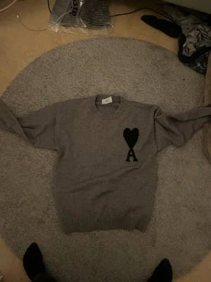 Grå stickad tröja från Ami - Snygg grå stickad tröja från Ami med rund halsringning och ribbade muddar. På bröstet finns ett svart hjärta med bokstaven A broderat. Perfekt för dig som gillar stilrena plagg med en twist. Nästan helt ny S men sitter som M. Lapp följer med 