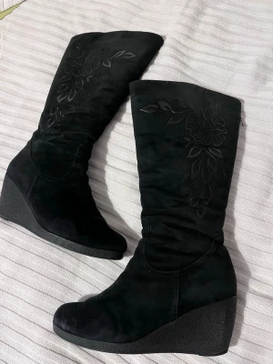 Svarta kilklackade stövlar med blommönster - Snygga svarta stövlar med kilklack och blommigt mönster på skaftet. Tillverkade i mjuk mocka som ger en lyxig känsla. Perfekta för dig som vill ha både höjd och stil i vardagen. Lätt att matcha med olika outfits. Passar bäst om du har storlek 39-39,5 Avdragare/Kläpp (Puller) fixade nyss, lite grövre än innan men de funkar.