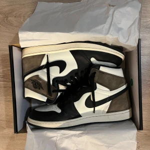 Nike Air Jordan 1 High Mocha - Nike Air Jordan 1 High Mocha med svart och vit läder, bruna mockadetaljer och klassisk svart swoosh. Storlek 47. Höga sneakers med snörning, perforerad tå och ikonisk Jordan-logga på sidan. Perfekt för dig som vill sticka ut med en clean och tidlös look.