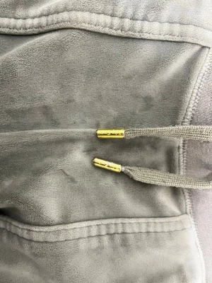 Grå velour mjukisbyxor med snörning - Snygga grå mjukisbyxor i mjuk velour med breda ben och guldfärgade detaljer på snöret. Byxorna har resår i midjan och två bakfickor med knapp. Perfekta för chill dagar eller när du vill vara extra comfy.