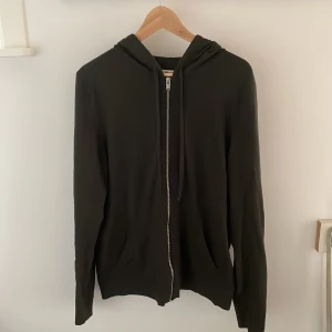 Olivgrön hoodie från Zadig & Voltaire - Olivgrön hoodie från Zadig & Voltaire med dragkedja framtill och huva med snörning. På ärmarna finns svarta blixtdetaljer och broderad ZV-logga. Tröjan är stickad i merinoull och har en avslappnad passform, perfekt för chill dagar.