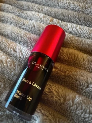 Clarins Water Lip Stain röd - Clarins Water Lip Stain i en djup röd nyans. Kommer i en smidig glasflaska med metallicrött lock och guldig text. Perfekt för dig som vill ha en lätt men intensiv färg på läpparna som sitter länge.