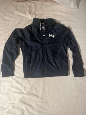 Marinblå vindjacka från Helly Hansen - Säljer en marinblå vindjacka från Helly Hansen med vit HH-logga på bröstet och ryggen. Jackan har hög krage, dragkedja framtill och två sidofickor med dragkedja. Tillverkad i polyester och har resår vid ärmslut och midja för skön passform.