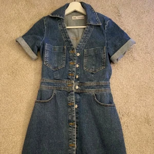 Jeansklänning med knappar från ZARA - Säljer en snygg jeansklänning från ZARA med korta ärmar, v-ringning och knappar hela vägen framtill. Klänningen har två bröstfickor och markerad midja för en schysst passform. Perfekt till sneakers eller boots!