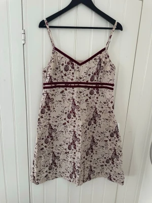 Blommig kortklänning med vinröda detaljer - Supersöt kortklänning med smala axelband och v-ringning. Klänningen är beige med vinrött blommönster och har två sammetsband under bysten. Dragkedja i sidan för enkel påklädning. Perfekt för sommarens alla tillfällen!