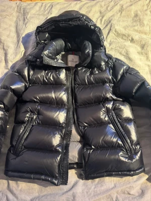 Svart pufferjacka från Moncler - Säljer en svart pufferjacka från Moncler med glansig finish och huva. Jackan har dragkedja framtill, två snedställda fickor med dragkedja och Moncler-logga på ärmen. Perfekt för kalla dagar och riktigt snygg till streetwear.