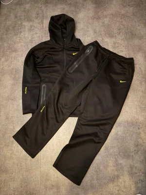 Svart NOCTA loungewear set från Nike - Snyggt svart loungewear set från Nike NOCTA med hoodie och matchande byxor. Setet har gula detaljer, dragkedjor på fickorna och NOCTA-tryck på ärm och ben. Perfekt för chill dagar hemma eller när du vill vara bekväm men ändå se fresh ut. PASSAR DIG SOM BRUKAR BÄRA L