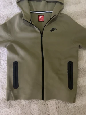 Olivgrön Nike hoodie med dragkedja - Snygg olivgrön hoodie från Nike med svart logga på bröstet och hel dragkedja framtill. Två svarta dragkedjeförsedda fickor och huva. Tillverkad i mjukt material, perfekt för en avslappnad streetwear-look.