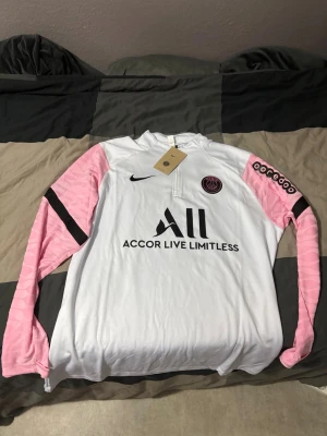 PSG träningsset från Nike, vit/rosa - Snyggt PSG träningsset från Nike med långärmad vit tröja, rosa och svarta detaljer på ärmarna samt PSG-logga och sponsortryck framtill. Matchande svarta byxor med rosa och svart detalj samt PSG-märke. Perfekt för fotbollsträning eller chill.
