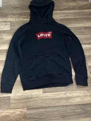 Marin blå hoodie från Levi's med logga - Säljer en marin blå hoodie från Levi's med den klassiska röda loggan på bröstet. Hoodien har huva, magficka och ribbade muddar vid ärmslut och nederkant. Perfekt för en avslappnad och cool stil. Materialet känns mjukt och bekvämt. Jätte fin och skön ! Storlek är S men passar även som xs