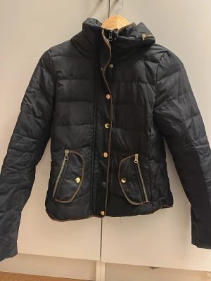 Svart quiltad pufferjacka med guldknappar - Snygg svart pufferjacka med quiltad design och guldfärgade knappar och dragkedjor. Jackan har hög krage, två framfickor med dragkedja och dekorativa detaljer i brunt. Perfekt för kyliga dagar och ger en trendig look.