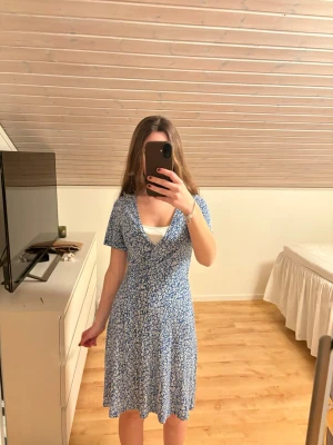 Blå blommig omlottklänning - Söt blå kortklänning med vitt blommönster. Klänningen har v-ringning och korta ärmar, perfekt för varma dagar. Materialet känns mjukt och lätt, troligen bomull eller polyester. Enkel och fräsch stil som passar till många tillfällen.