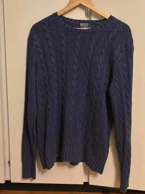 Mörkblå kabelstickad tröja Polo Ralph Lauren - Kabelstickad mörkblå tröja från Polo Ralph Lauren. Klassisk rund halsringning och ribbade muddar vid ärmslut och nederkant. Perfekt för lager på lager och en tidlös stil till garderoben.