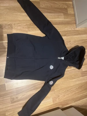 Svart hoodie från Moncler med dragkedja - Säljer en svart hoodie från Moncler med dragkedja framtill och två fickor. Hoodien har Moncler-logga på bröstet och ärmen samt ribbade muddar. Tillverkad i mjukt bomullsmaterial och har en klassisk huva. Perfekt för en avslappnad och stilren look.
