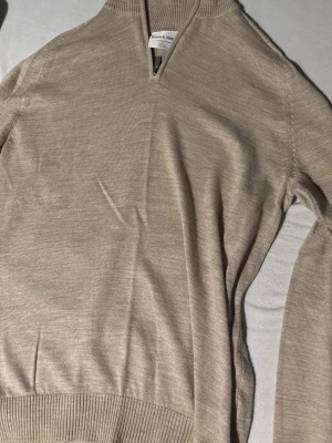 Beige stickad tröja från Bruun & Stengade - Beige stickad tröja från Bruun & Stengade med halv dragkedja vid halsen och ribbade muddar. Tröjan har en stilren och enkel design, perfekt för lager-på-lager och passar till många olika outfits.