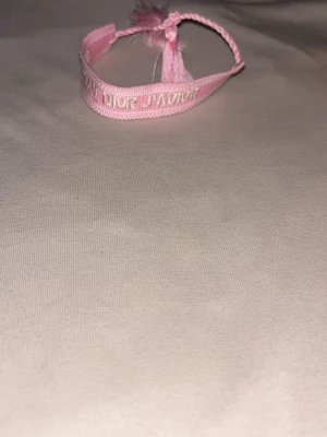 Rosa J'Adior armband från Dior - Säljer ett rosa tygarmband från Dior med texten 'Christian Dior J'Adior' broderad i vitt. Armbandet har justerbar knytning och tofsar i ändarna. Perfekt accessoar för att lyfta din stil med en lyxig touch.