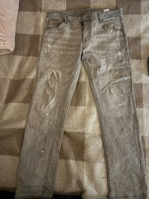 DSQUARED2slitna jeans med färgstänk - Säljer ett par grå jeans med vita färgstänk på både fram- och baksida. Jeansen har klassisk femficksdesign och raka ben. Perfekta för dig som vill ha en unik och trendig look.