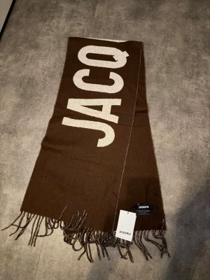 Brun halsduk från Jacquemus - Snygg brun halsduk från Jacquemus med stora vita bokstäver och fransar i kanterna. Tillverkad i mjuk ullblandning och har en rektangulär form. Perfekt accessoar för att lyfta din outfit med en trendig touch.