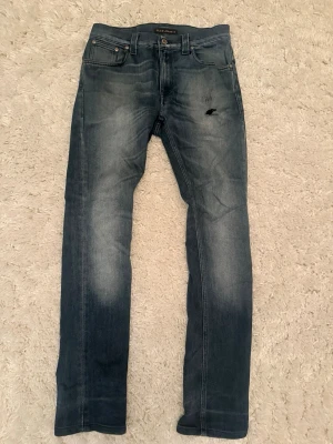Blå jeans från Nudie Jeans - Säljer ett par klassiska blå jeans från Nudie Jeans med raka ben och snygga slitningar. Jeansen har fem fickor, knappgylf och Nudie-loggan broderad på bakfickorna. Perfekta för dig som gillar en avslappnad och tidlös stil.