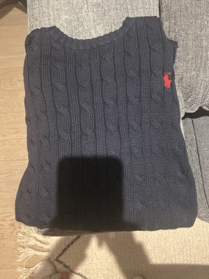 Mörkblå kabelstickad tröja Polo Ralph Lauren - Snygg mörkblå kabelstickad tröja från Polo Ralph Lauren med klassisk röd logga broderad på bröstet. Tröjan har rund halsringning och är gjord i mjukt stickat material. Perfekt för en stilren och avslappnad look.