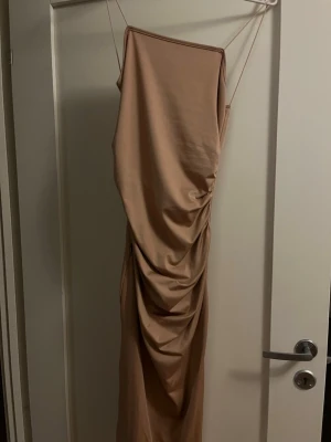 Beige långklänning med drapering - Säljer en beige långklänning med tunna axelband och snygg drapering längs sidan. Klänningen har en elegant, figurnära passform och är tillverkad i ett mjukt, stretchigt material som ger en lyxig känsla. Perfekt för dig som vill sticka ut med en stilren look.