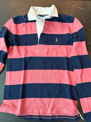 Randig rugbytröja från Ralph Lauren Sport - Klassisk rugbytröja från Ralph Lauren med breda marinblå och rosa ränder, vit krage och knappslå. Tröjan har lång ärm och ikonisk broderad logga på bröstet. Perfekt för en sportig och avslappnad stil.