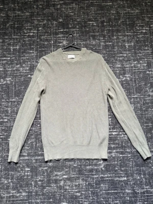 Olivgrön stickad tröja med rund hals - En stilren ljusgrå stickad tröja med rund hals och långa ärmar. Tröjan har ribbade muddar vid ärmslut och nederkant, vilket ger en snygg passform. Perfekt för lager på lager och enkel att matcha med olika outfits.