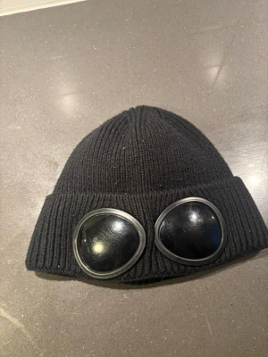 Svartgoggle mössa - Säljer en svart ribbstickad mössa med de ikoniska goggle-linserna framtill. Mössan är tillverkad i mjukt material och har en uppvikt kant. Perfekt för dig som vill sticka ut med streetwear-stil.
