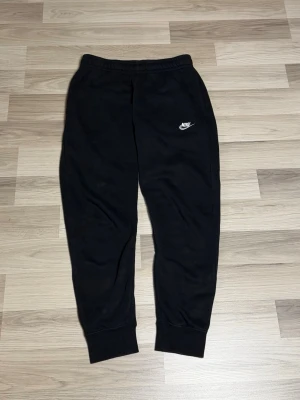 Svarta mjukisbyxor från Nike - Svarta mjukisbyxor från Nike med klassisk logga på vänster lår. Byxorna har resår i midjan och muddar vid bensluten för en skön och avslappnad passform. Perfekta för chill eller träning. Tillverkade i mjukt bomullsmaterial.