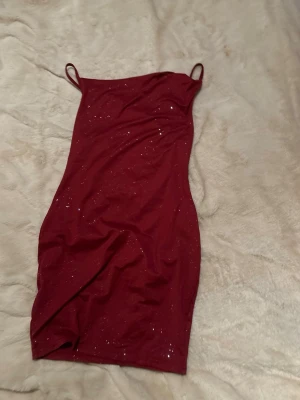 Vinröd glittrig kort klänning - Snygg vinröd kort klänning med tunna axelband och glittriga detaljer. Klänningen har en figurnära passform och är perfekt för dig som vill sticka ut. Materialet känns mjukt och stretchigt, vilket gör den bekväm att bära hela kvällen.