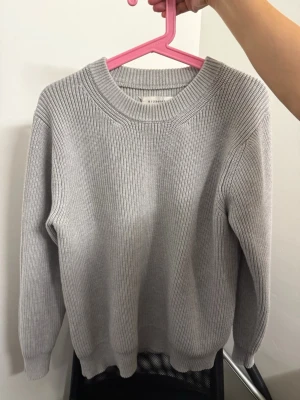 Maison Margeila Grey knit - En stilren ljusgrå stickad tröja med ribbstickad struktur och rund hals. Tröjan har långa ärmar och breda muddar vid ärmslut och nederkant. Perfekt för lager på lager och enkel att matcha med olika outfits.