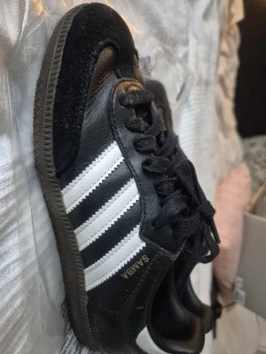 Adidas Samba svarta sneakers stl 37 1/3 - Klassiska Adidas Samba sneakers i svart skinn med vita ränder på sidorna och svart mockadetalj vid tån. Skon har snörning och en brun gummisula med mönster undertill. Perfekt för dig som gillar retrostil och sportig look. Storlek 37 1/3.
