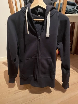 Russemerch tröja - Mörkblå hoodie med dragkedja från Russemerch. Har stora fickor framtill, vit dragsko i huvan och ribbade muddar vid ärmslut och nederkant. Tryck med Russedress på ena ärmen. Perfekt för chill dagar eller när du vill ha en enkel och snygg look.