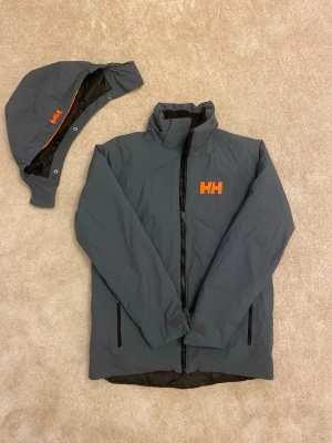 Helly Hansen skidjacka – stl 164 (14 år) - Snygg och funktionell skidjacka från Helly Hansen. Den är mörkgrå med oranga detaljer. Jackan är sparsamt använd och i väldigt bra skick.  Den är varm, vind- och vattenavvisande- perfekt för skidåkning etc. Avtagbar huva som enkelt knäpps vid behov.   Detaljer •	Storlek: 164 / 14 år •	Märke: Helly Hansen •	3 fickor med dragkedja (en på ärmen för liftkort) •	Avtagbar huva •	Justerbara ärmslut •	Sparsamt använd, inga kända skador eller slitage