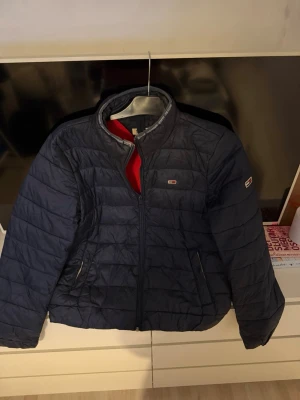 Mörkblå dunjacka från Tommy Jeans - Snygg mörkblå dunjacka från Tommy Jeans med quiltad design och dragkedja framtill. Jackan har diskreta logodetaljer på bröstet och ärmen samt två fickor med knapp. Ordpris:2300kr säljer för 599kr. Storlek L.