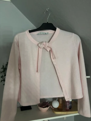 Ljusrosa kofta med knytband - Söt ljusrosa kofta från Tina med långa ärmar och knytband framtill. Koftan har en rund halsringning och är gjord i ett mjukt, tunt material som känns skönt mot huden. Perfekt att slänga över en topp för en extra touch.