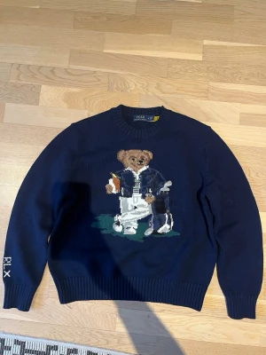 Mörkblå Polo Ralph Lauren björntröja - Nästintill helt ny stickad mörkblå tröja från Polo Ralph Lauren med ikonisk björn som håller en drink och golfklubbor på bröstet. Rund halsringning och RLX-brodyr på ärmen. Använd 1 gång. Passar ca 170 cm. 