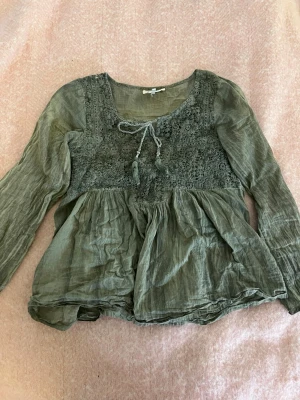 Olivgrön boho-blus med spetsdetaljer Stockholmsstil - Säljer en olivgrön boho-inspirerad blus med långa ärmar och vackra spetsdetaljer över bröstet. Blusen har en lös passform, knytband med tofsar vid halsen och är tillverkad i 100% bomull som ger en avslappnad look.