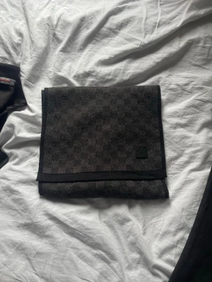Mörkgrå rutig halsduk Louis Vuitton - Snygg mörkgrå halsduk från Louis Vuitton med diskret rutmönster och svart kant. Tillverkad i ett mjukt material som känns lyxigt och stilrent. Perfekt accessoar för att lyfta din outfit.