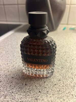 Valentino parfym med nitar och mönster - Säljer en lyxig parfym från Valentino i en genomskinlig glasflaska med nitar och rutmönster. Flaskan har svart kork och etikett med Valentino-loggan i rött. Innehållet är bärnstensfärgat och flaskan har en exklusiv och edgy design.