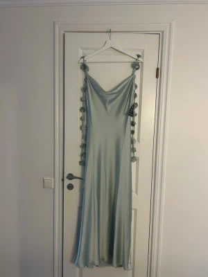 Ljusblå långklänning med knytband - Säljer en elegant ljusgrön långklänning i satinliknande material. Klänningen har tunna axelband med dekorativa knyt och en snygg draperad urringning. Perfekt för dig som vill ha en stilren och trendig look.