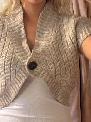 Beige stickad bolero med knapp - Kort beige stickad bolero från Gina Tricot med bred ribbad krage och ett stort dekorativt knappfäste framtill. Modellen har korta ärmar och ett diskret mönster i stickningen. Jag är 180cm lång och bär vanligtvis S/M. Skicka gärna frågor eller prisförslag!