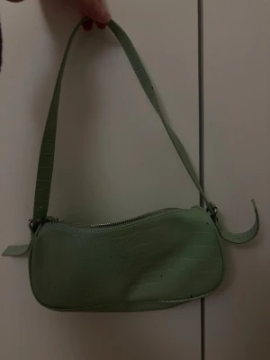 Grön handväska med dragkedja - Snygg ljusgrön handväska i syntetmaterial med croco-mönster. Väskan har en enkel axelrem och stängs med dragkedja upptill. Perfekt för dig som vill ha en trendig accessoar med stilren look. Lite sliten men ser annars ny ut.