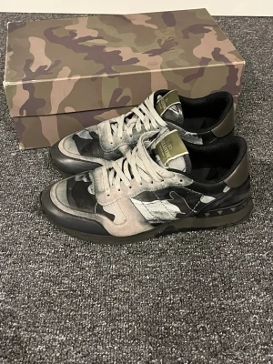 Valentino Rockrunner - Tja jag säljer ett par Valentino Rockrunner sneakers med grå, svart och gråvit camouflage-mönster. Skorna har snörning, rund tå och detaljer i mocka och skinn. Snygg och unik design med klassiska Valentino-nitar på sulan. Box medföljer. Kom gärna med frågor om det är några funderingar. Priset är ej hugget i sten mvh Hugo