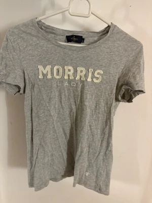 Grå t-shirt från Morris Lady - Grå t-shirt från Morris Lady med broderad vit text på bröstet. Klassisk rund halsringning och korta ärmar. Tillverkad i mjuk bomull som känns skön mot huden. Perfekt för en avslappnad och stilren look.