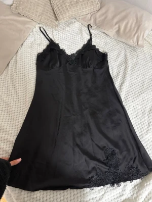 Svart nattlinne med spetsdetaljer - Snyggt svart nattlinne i satinliknande material med tunna axelband och v-ringning. Dekorerat med spetsdetaljer vid bysten och längs nederkanten, vilket ger en elegant och lyxig känsla. Perfekt för mysiga kvällar hemma.