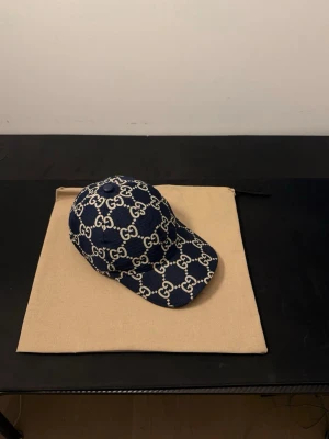 Blå Gucci keps med monogram - Snygg blå keps från Gucci med det klassiska GG-monogrammet i vitt över hela kepsen. Tillverkad i ett slitstarkt tyg med böjd skärm och justerbar baksida. Perfekt för dig som vill ha en trendig accessoar med lyxig känsla. Pris kan diskuteras!!