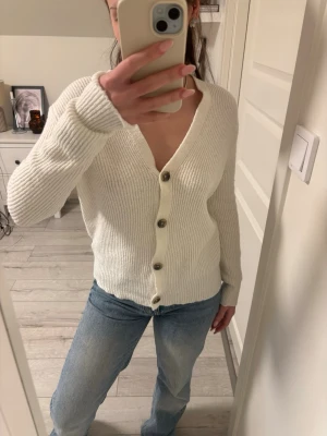 Vit ribbad kofta med knappar - Säljer en stilren vit ribbad kofta med v-ringning och stora knappar framtill. Koftan har lång ärm och är gjord i ett mjukt stickat material som känns skönt mot huden. Perfekt att slänga över en topp eller bära som den är.