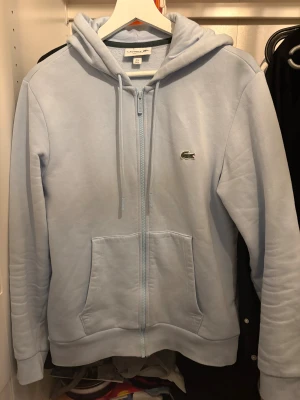 Ljusblå hoodie från Lacoste - Snygg ljusblå hoodie från Lacoste med klassisk krokodillogga på bröstet. 